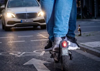 Rückansicht von zwei Personen, die sich einen E-Scooter auf einem Radweg mit Fahrradsymbol teilen, während sich im Hintergrund ein Taxi bei Dämmerung nähert.