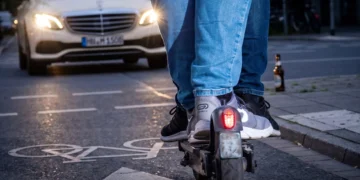 Rückansicht von zwei Personen, die sich einen E-Scooter auf einem Radweg mit Fahrradsymbol teilen, während sich im Hintergrund ein Taxi bei Dämmerung nähert.