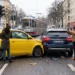 Frontalzusammenstoß zwischen einem gelben Kleinwagen und einem dunklen SUV an einer Straßenkreuzung, Beteiligte und Zeugen stehen daneben.