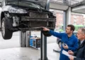 Kfz-Mechaniker zeigt einem Kunden in der Werkstatt die freiliegende Front eines auf der Hebebühne stehenden Fahrzeugs mit demontierter Stoßstange.