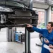 Kfz-Mechaniker zeigt einem Kunden in der Werkstatt die freiliegende Front eines auf der Hebebühne stehenden Fahrzeugs mit demontierter Stoßstange.