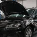 Kfz-Mechaniker poliert die beschädigte Seitenwand eines schwarzen VW mit geöffneter Motorhaube in einer Autowerkstatt.