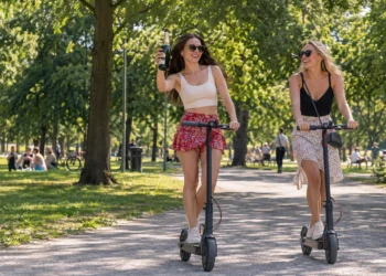 Zwei Frauen fahren im Sommer mit E-Scootern durch einen Park, eine hält eine offene Sektflasche in der Hand.