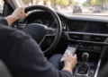 Blick vom Beifahrersitz auf einen Autofahrer, der die linke Hand am Lenkrad hat und mit der rechten Hand ein Smartphone über der Mittelkonsole hält und bedient, während vor dem Fahrzeug dichter Stadtverkehr zu sehen ist.