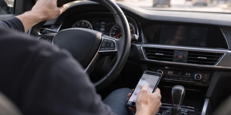 Blick vom Beifahrersitz auf einen Autofahrer, der die linke Hand am Lenkrad hat und mit der rechten Hand ein Smartphone über der Mittelkonsole hält und bedient, während vor dem Fahrzeug dichter Stadtverkehr zu sehen ist.