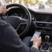 Blick vom Beifahrersitz auf einen Autofahrer, der die linke Hand am Lenkrad hat und mit der rechten Hand ein Smartphone über der Mittelkonsole hält und bedient, während vor dem Fahrzeug dichter Stadtverkehr zu sehen ist.