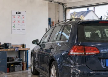 Dunkler Kombi mit eingedrückter Seitenwand steht in einer Garage; an der Wand hängt ein Kalender, auf dem sechs Monate durchgestrichen sind und im siebten ein Euro-Zeichen steht.
