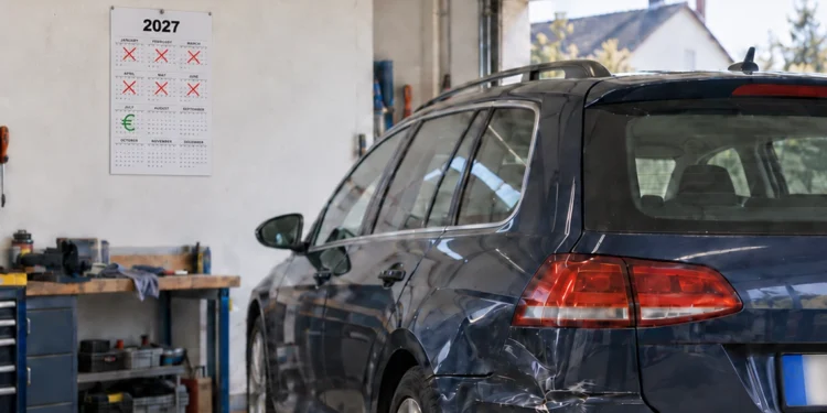 Dunkler Kombi mit eingedrückter Seitenwand steht in einer Garage; an der Wand hängt ein Kalender, auf dem sechs Monate durchgestrichen sind und im siebten ein Euro-Zeichen steht.