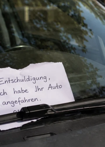 Handgeschriebener Zettel nach einem Parkrempler unter dem Scheibenwischer eines geparkten Autos, mit teilweise verdeckter und nicht lesbarer Telefonnummer