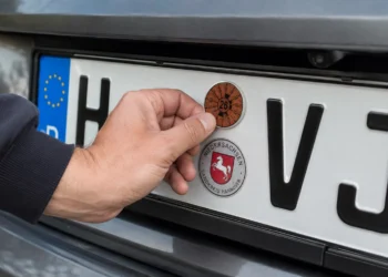 Hand befestigt eine braune HU-Plakette für 2026 auf einem Kennzeichen mit dem Kürzel H‑VJ, darunter das Niedersachsen-Siegel