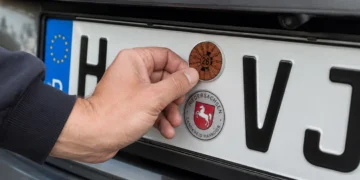 Hand befestigt eine braune HU-Plakette für 2026 auf einem Kennzeichen mit dem Kürzel H‑VJ, darunter das Niedersachsen-Siegel