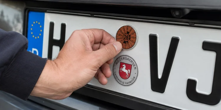 Hand befestigt eine braune HU-Plakette für 2026 auf einem Kennzeichen mit dem Kürzel H‑VJ, darunter das Niedersachsen-Siegel
