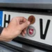 Hand befestigt eine braune HU-Plakette für 2026 auf einem Kennzeichen mit dem Kürzel H‑VJ, darunter das Niedersachsen-Siegel