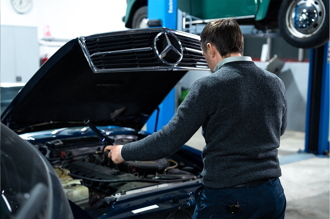 Ilya Bogdanovskiy von hinten über dem geöffneten Motorraum eines älteren Mercedes in einer Werkstatt, dahinter ein Fahrzeug auf der Hebebühne.