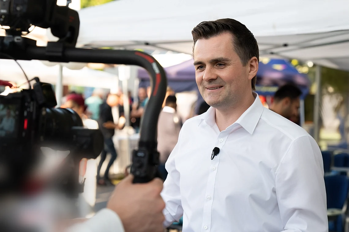 Ilya Bogdanovskiy steht im Freien vor einer Filmkamera auf einem Gimbal und spricht ins Bild, im Hintergrund Zelte und Besucher einer Veranstaltung.