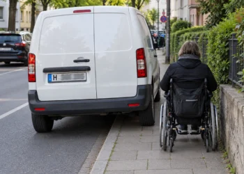 Weißer Transporter parkt quer auf dem Gehweg, eine Rollstuhlfahrerin muss auf den schmalen Rest ausweichen