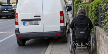 Weißer Transporter parkt quer auf dem Gehweg, eine Rollstuhlfahrerin muss auf den schmalen Rest ausweichen
