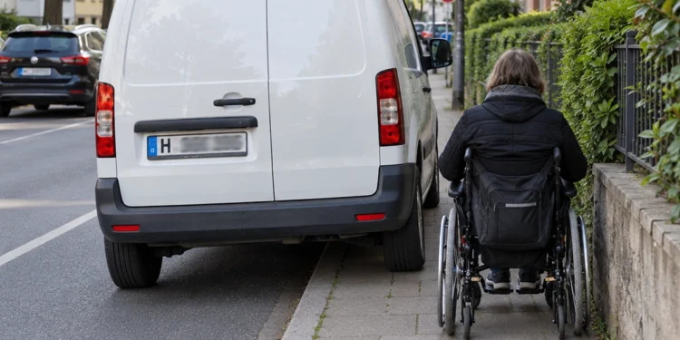 Weißer Transporter parkt quer auf dem Gehweg, eine Rollstuhlfahrerin muss auf den schmalen Rest ausweichen