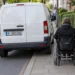 Weißer Transporter parkt quer auf dem Gehweg, eine Rollstuhlfahrerin muss auf den schmalen Rest ausweichen