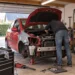 Roter Kleinwagen mit abgebauter Front steht auf Unterstellböcken in einer privaten Garage, ein Mann arbeitet über dem offenen Motorraum, daneben Werkzeugwagen und Wagenheber.