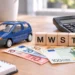 Blaues Modellauto neben Holzwürfeln mit der Aufschrift MWST, Euro-Banknoten und einem Taschenrechner auf einem Schreibtisch