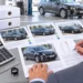 Schreibtisch mit ausgedruckten Vergleichsangeboten mehrerer gleichartiger Kombis, daneben ein Taschenrechner mit der Anzeige 130 %, im Hintergrund ein Unfallwagen in der Werkstatt.