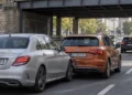 Unter einer Bahnbrücke im Stadtverkehr ist eine silberne Limousine auf das Heck eines orangefarbenen Kompaktwagens aufgefahren, die hintere Stoßstange ist eingedrückt.
