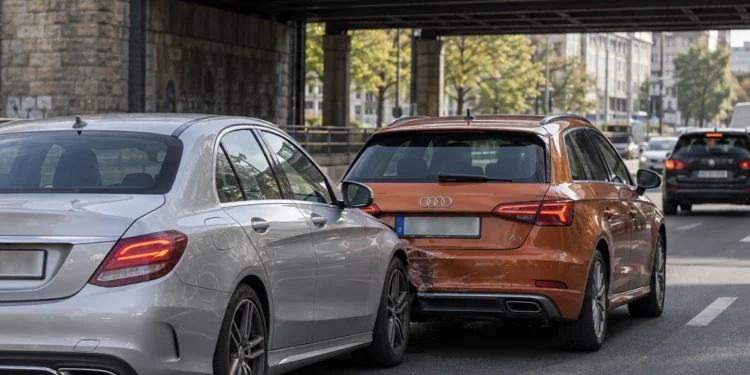 Unter einer Bahnbrücke im Stadtverkehr ist eine silberne Limousine auf das Heck eines orangefarbenen Kompaktwagens aufgefahren, die hintere Stoßstange ist eingedrückt.