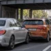 Unter einer Bahnbrücke im Stadtverkehr ist eine silberne Limousine auf das Heck eines orangefarbenen Kompaktwagens aufgefahren, die hintere Stoßstange ist eingedrückt.