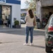 Eine Frau steht mit dem Handy am Ohr zwischen einer großen Markenwerkstatt und einer kleinen freien Werkstatt, rechts parkt ein dunkelgrünes Auto mit Heckschaden.