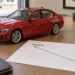 Ein rotes Modellauto einer Limousine steht auf einem Schreibtisch neben einem Blatt mit einer fallenden Wertlinie von 45.000 auf 30.000 Euro.