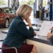 Eine Kundin sitzt am Tresen einer Werkstatt und schaut nachdenklich auf eine Reparaturrechnung, die ihr ein Mitarbeiter zeigt, im Hintergrund ein braunes Auto auf der Hebebühne.